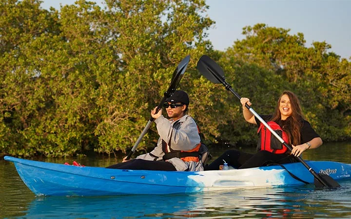 Two people, a man and a woman, paddle a blue kayak together on calm water under the glow of Full Moon Kayaking Qatar, surrounded by green trees and sunlight. Both wear life jackets and appear to be enjoying themselves.