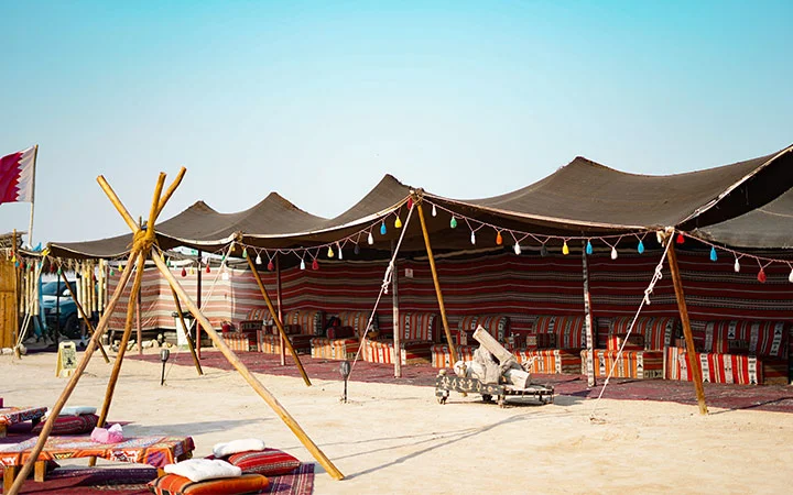 A traditional Bedouin-style tent with colorful cushions and rugs is set up on sandy ground under a clear blue sky, evoking the spirit of Full Moon Kayaking Qatar. Multi-colored lights hang along the tent’s edge and wooden poles stand in the foreground.