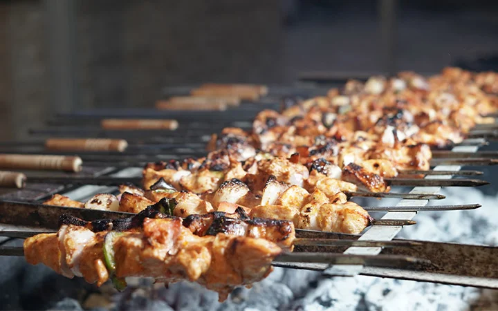 Skewers of marinated meat and vegetables are grilling over hot charcoal during the Full Moon Desert Safari, with a smoky, charred texture visible on the food and wooden handles extending out from the grill.