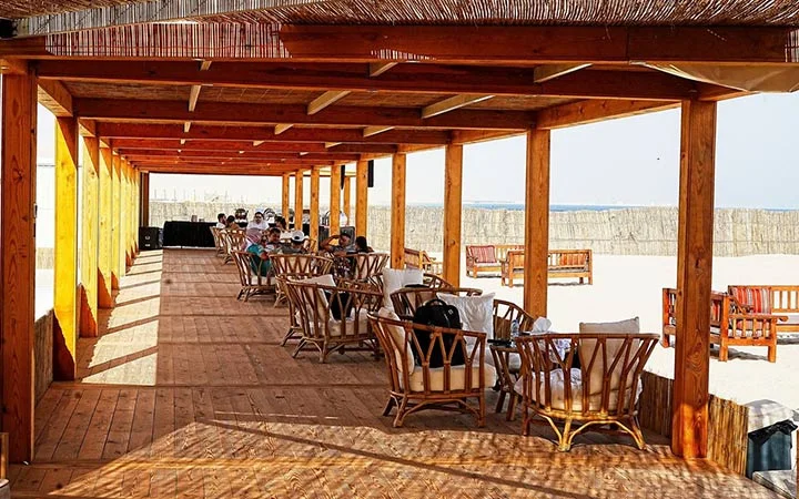People sit and relax on wooden chairs under a shaded beachside pavilion with wooden beams, overlooking a sandy area and the sea—an ideal spot to unwind after a Full Moon Desert Safari.