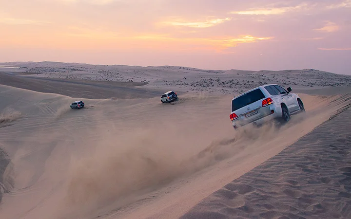 Three SUVs drive up and across sandy dunes at sunset on a Full Moon Desert Safari, kicking up clouds of sand behind them under a colorful sky in a vast desert landscape.