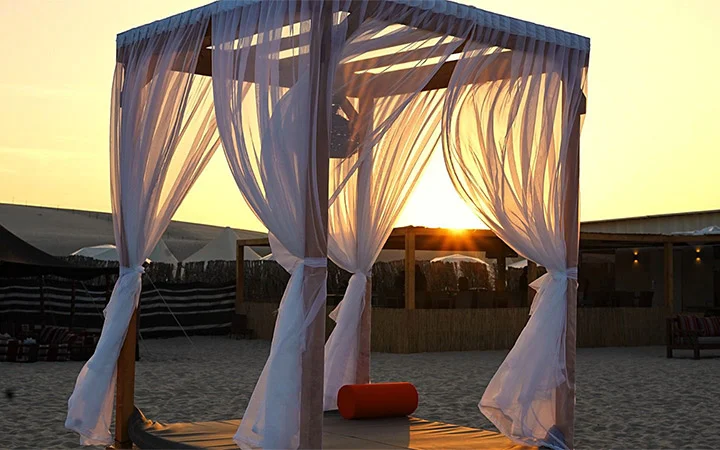 A cabana with sheer white curtains tied to wooden posts stands on a sandy beach at sunset, bathed in the warm light—evoking the tranquility of a Full Moon Desert Safari.