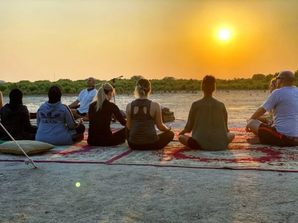 A group of people sit cross-legged on mats outdoors at sunset, facing an instructor, enjoying a relaxing meditation or yoga session before joining a Treasure Hunt on Purple Island, with a stunning natural landscape in the background.