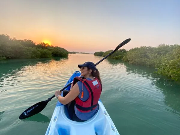 A woman wearing a life jacket and cap enjoys Mangrove Kayaking in Qatar, gliding on calm water at sunset surrounded by lush green mangroves, smiling as she looks back toward the camera.