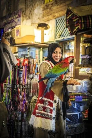 A smiling woman in a hijab and patterned vest holds a colorful parrot in a busy indoor market filled with pet supplies—like she's on a Treasure Hunt on Purple Island, discovering vibrant surprises at every turn.