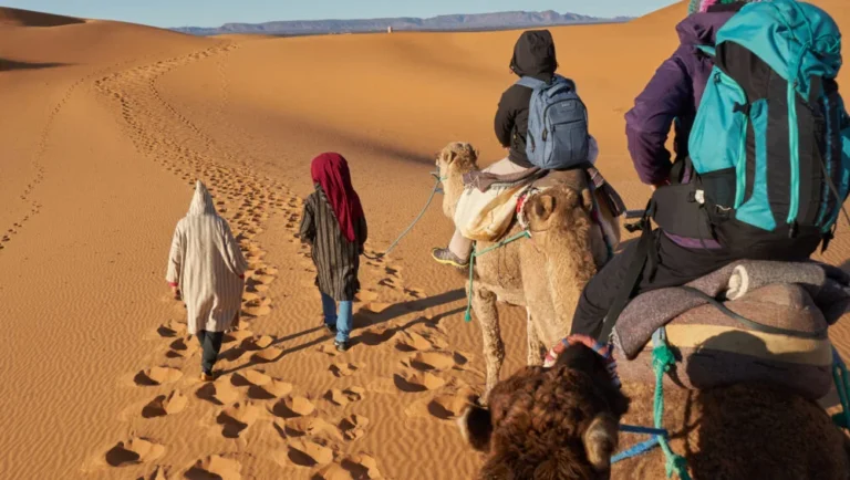 A group of people ride camels led by two guides walking through sandy desert dunes, exploring the landscape with Qatar desert safari packages, leaving footprints and hoofprints behind them under a clear sky.