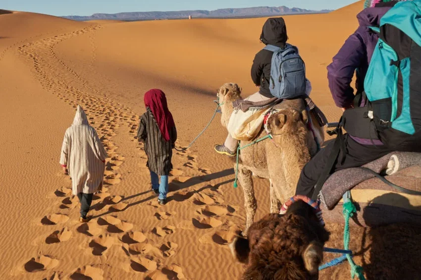 A group of people ride camels led by two guides walking through sandy desert dunes, exploring the landscape with Qatar desert safari packages, leaving footprints and hoofprints behind them under a clear sky.