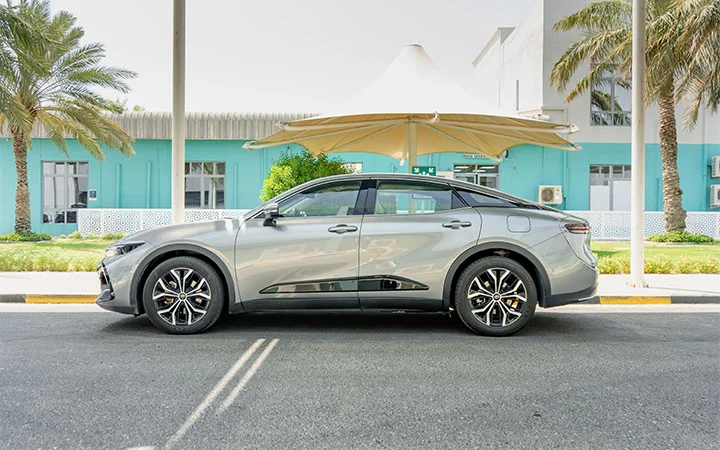 A silver modern electric car, resembling the sleek style of a Toyota Camry Qatar, is parked on a sunny street with palm trees and a turquoise building in the background. The car is viewed from the side, under a white canopy.