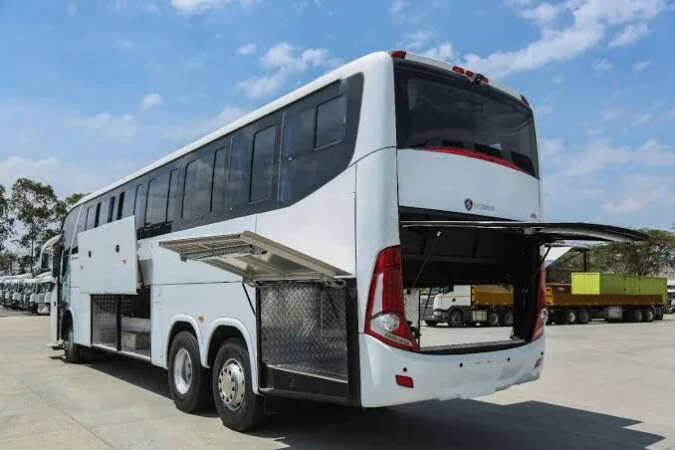 A large white VIP Coach for Driver Qatar is parked with its storage compartments and rear hatch open, revealing ample luggage space. Several trucks are visible in the background under a partly cloudy sky.