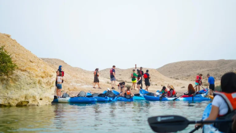A group of people in life jackets stand and sit on blue kayaks by a rocky shoreline at Purple Island Qatar, with some on land and others in the water, under a clear sky in a desert-like landscape.
