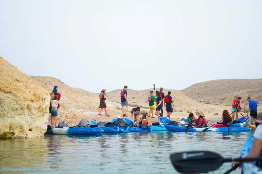 A group of people in life jackets stand and sit on blue kayaks by a rocky shoreline at Purple Island Qatar, with some on land and others in the water, under a clear sky in a desert-like landscape.
