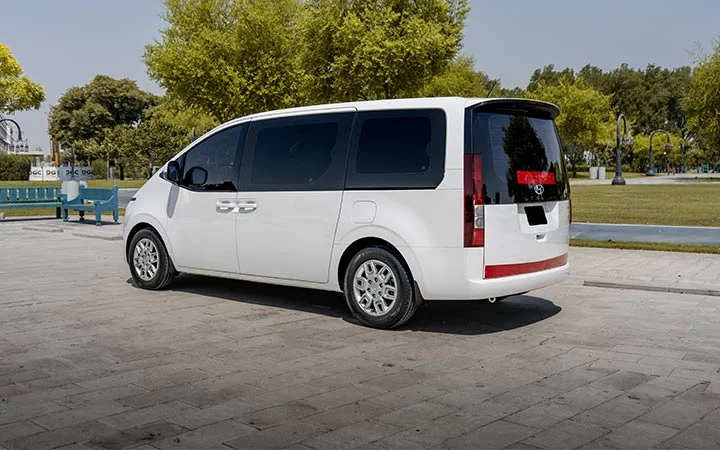 A white minivan from Mini Bus Vans Qatar is parked on a paved area near a park, with green trees and a blue bench in the background on a sunny day.