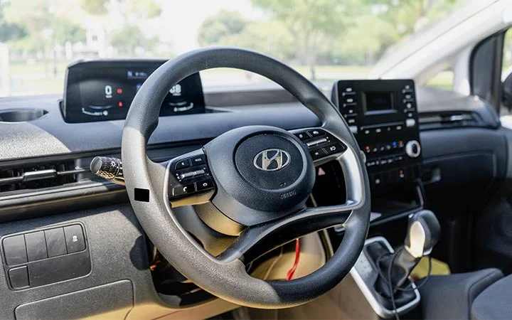Close-up view of a Hyundai car interior, showing the steering wheel with the Hyundai logo, dashboard screens, center console controls, and gear shift—ideal inspiration for Mini Bus Vans Qatar interiors. Blurred greenery is visible through the windows.