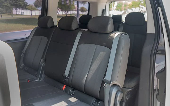 Interior view of a Mini Bus Vans Qatar showing two rows of gray and black fabric seats with headrests and seat belts; the side door is open and trees are visible outside.