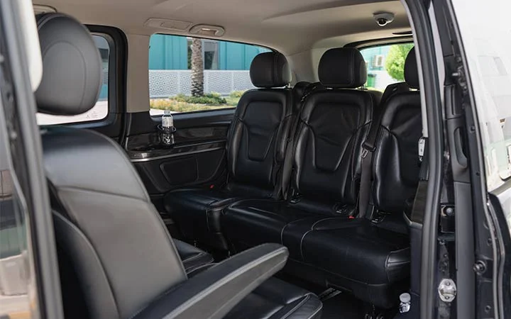 The interior of a Mercedes Viano in Qatar features black leather seats arranged in two rows. The sliding door is open, revealing spacious seating and a bottle of water on a side shelf, with sunlight streaming through the windows.