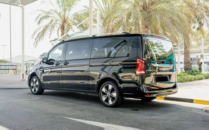A black Mercedes Viano in Qatar is parked on a street under a canopy, with palm trees and buildings visible in the background on a sunny day.