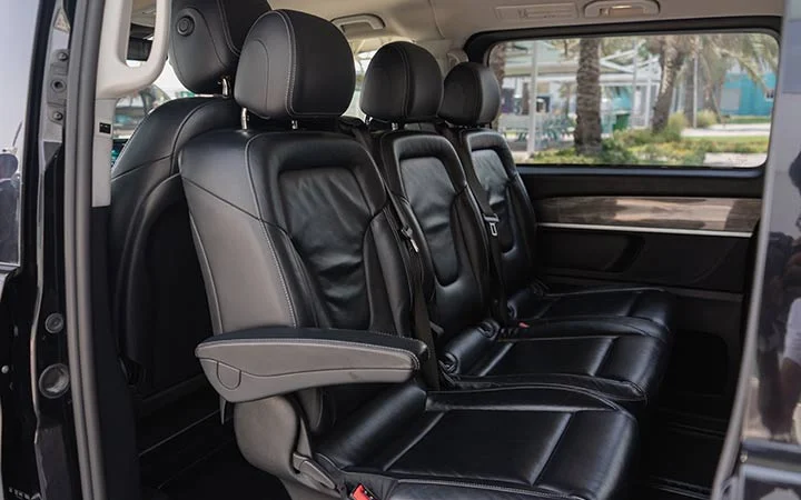 The image shows the interior of a Mercedes Viano in Qatar, featuring two rows of black leather seats with armrests, headrests, and seat belts. Large windows reveal palm trees and buildings outside.