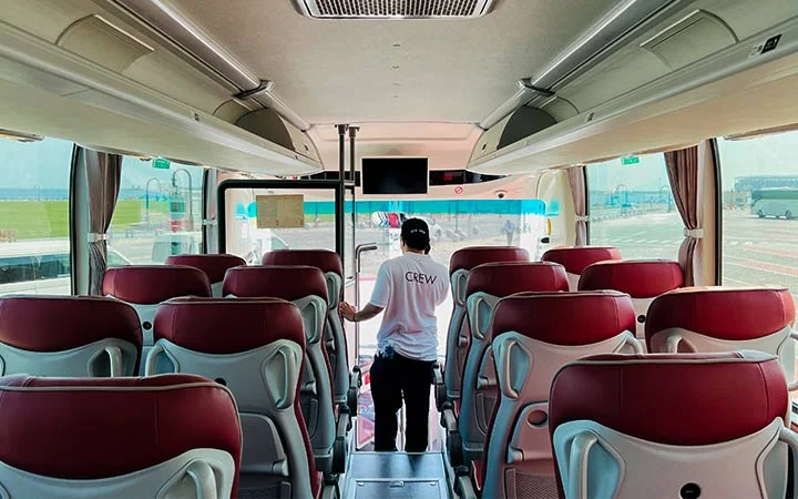 A person wearing a CREW shirt stands near the front of a Premium Coach Bus Qatar with red and gray seats, facing the open door and bright sunlight outside.