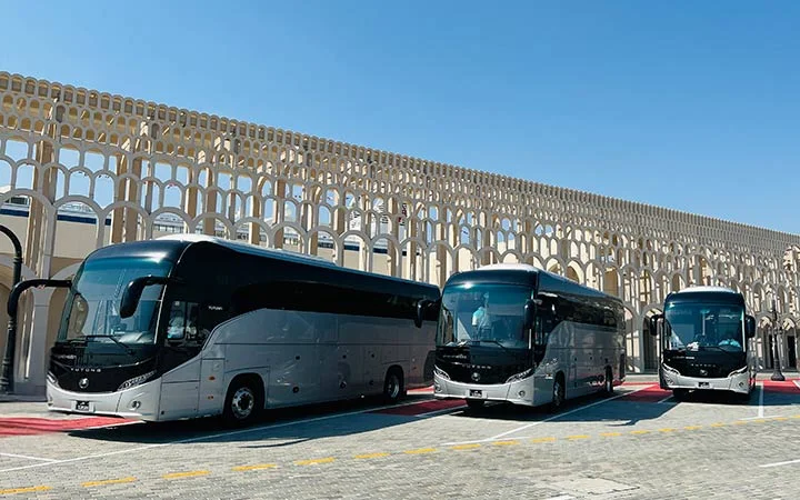 Three modern silver and black Premium Coach Bus Qatar vehicles are parked in a row on a paved area in front of a decorative arched building under a clear blue sky.