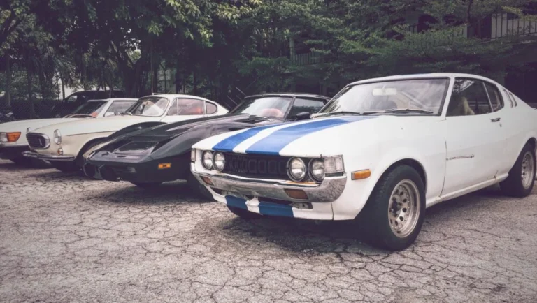 Four classic cars, embodying automotive excellence at GIMS Qatar, are parked side by side on cracked pavement; the front is a white coupe with blue racing stripes, while the others—black and cream vintage sports cars—are surrounded by trees.