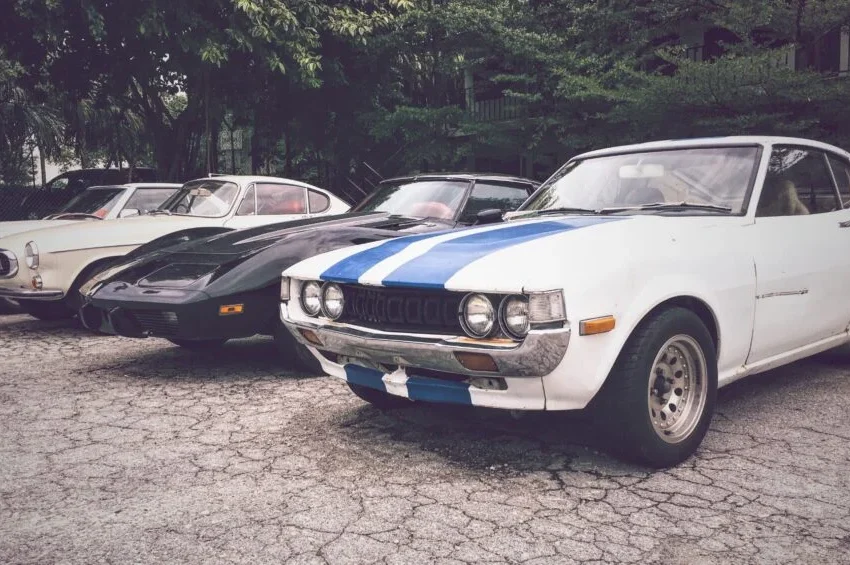 Four classic cars, embodying automotive excellence at GIMS Qatar, are parked side by side on cracked pavement; the front is a white coupe with blue racing stripes, while the others—black and cream vintage sports cars—are surrounded by trees.
