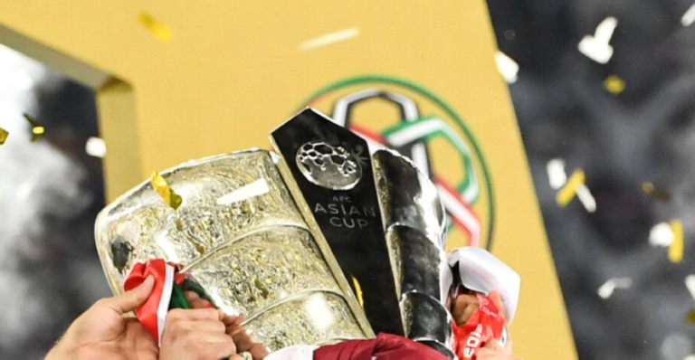 Close-up of hands holding the AFC Asian Cup trophy at afc qatar 2024, with confetti falling in the background and part of a large yellow structure visible behind the trophy.