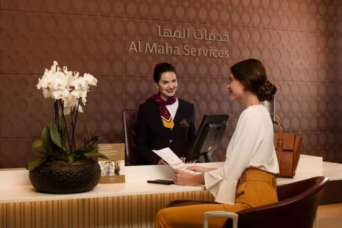 A female receptionist in uniform assists a smiling woman at the Al Maha Services desk, which features a brown patterned wall, a white orchid, brochures on the counter, and friendly support for al maha booking.