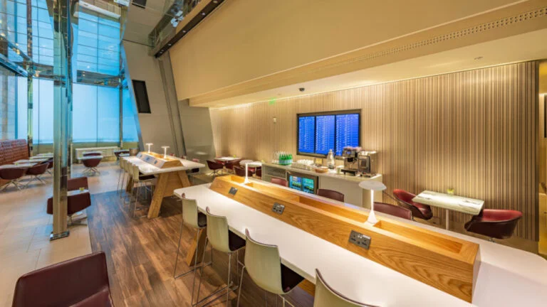 Modern airport lounge with wooden tables, white chairs, purple armchairs, and floor-to-ceiling windows. Enjoy al maha gold service perks near the digital flight information board and self-serve refreshment area in the warmly lit space.
