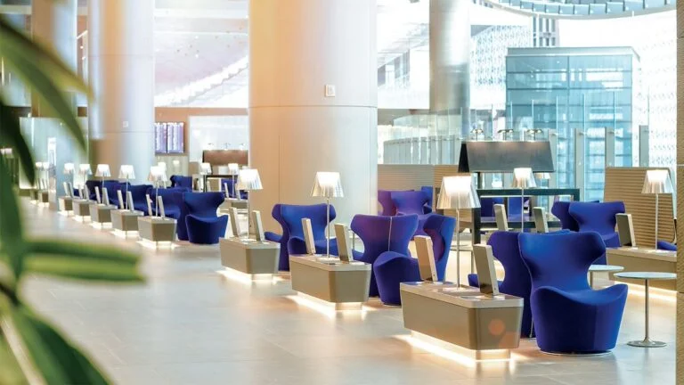 Modern airport lounge with rows of blue armchairs, individual lamp-lit tables, and large floor-to-ceiling windows. Enjoy a bright, spacious atmosphere with sleek design elements and the premium touch of Al Maha Service.