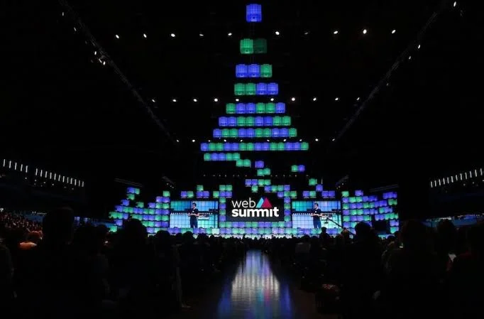 A large audience watches a stage at Web Summit Qatar 2024, with colorful blue and green illuminated blocks stacked in a pyramid shape above the stage and web summit displayed at the center.