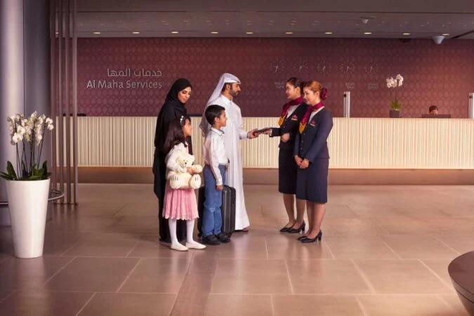 A family of four, including two children, is greeted by three airline staff in uniforms at an Al Maha Services check-in area, where a sign for al maha booking is visible. White orchids are placed in a pot by the entrance.
