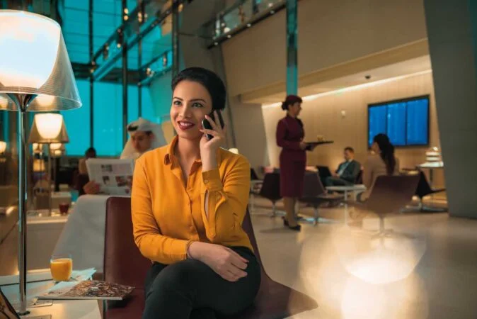 A woman in a yellow blouse sits in an airport lounge, smiling while talking on her phone. People are seated and a server offers al maha service in the background. A drink and magazine rest on the table beside her.
