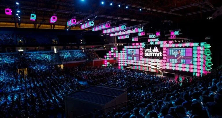 A large audience sits in a dimly lit arena, watching speakers on a brightly illuminated stage with digital screens displaying Web Summit Qatar 2024. Colorful pink and blue lights decorate the vibrant venue.