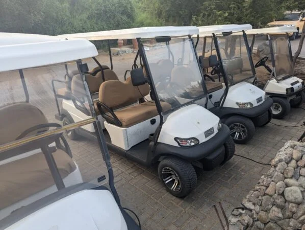Four white golf carts with beige seats are parked side by side on a paved area near a stone border and lush greenery, ready for your Al Maha booking adventure.