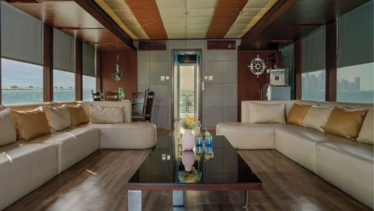 Modern yacht interior with large cream sofas, gold cushions, a black coffee table, and nautical anchors on the walls—perfect for Luxury Yacht Rentals—with expansive windows showcasing water views and the city skyline in the background.