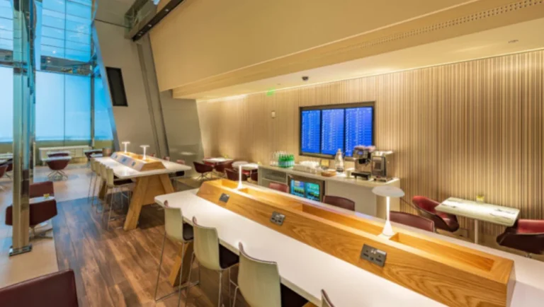 Modern airport lounge featuring Al Maha Gold Service by Qatar Airways, with wooden tables, white chairs, table lamps, and charging outlets. Enjoy a coffee station, snacks, and a large digital flight board set against wood-panel walls and bright windows.