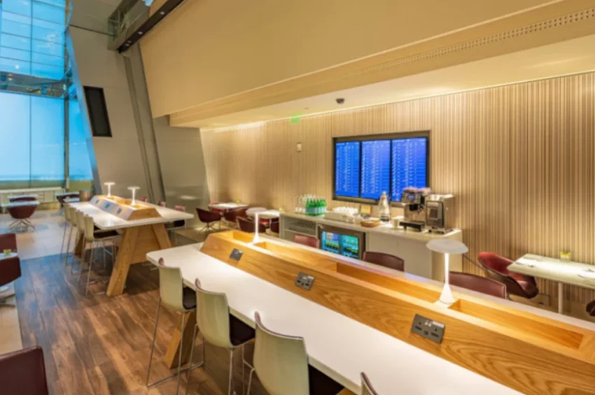 Modern airport lounge featuring Al Maha Gold Service by Qatar Airways, with wooden tables, white chairs, table lamps, and charging outlets. Enjoy a coffee station, snacks, and a large digital flight board set against wood-panel walls and bright windows.