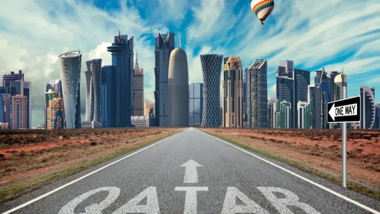 A straight road with QATAR and an arrow painted on it leads toward a modern city skyline—one of the top places to visit in Qatar. A hot air balloon floats in the blue sky, and a one-way street sign appears on the right side of the road.