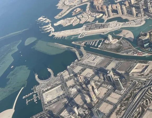Aerial view of Qatar’s coastal city with modern high-rises, artificial islands, marinas, and waterways surrounded by blue-green sea. Captured from a Piper Archer Plane, the urban layout contrasts with patches of undeveloped sandy land.