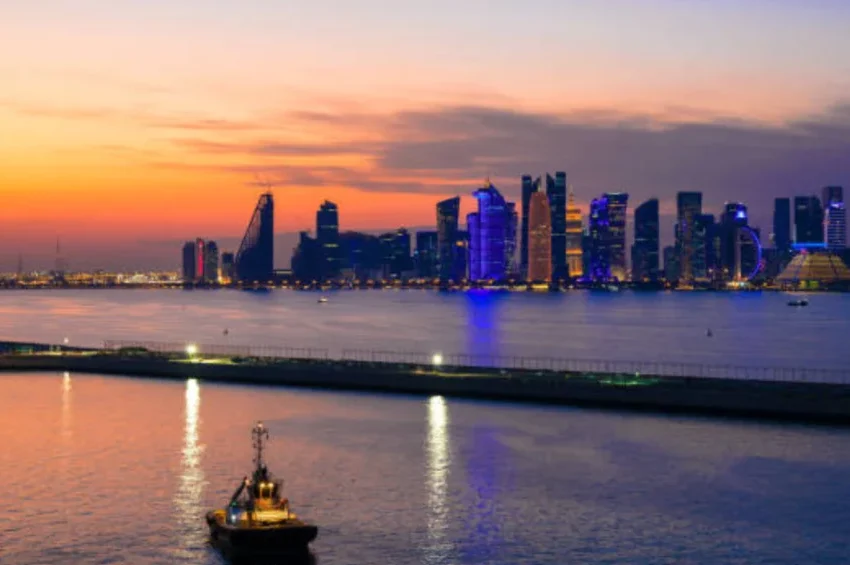 A small boat floats on calm water at sunset, with a brightly lit modern city skyline and tall skyscrapers in the background. The orange and purple sky reflects off the water, capturing the essence of the Best Sunset in Qatar.