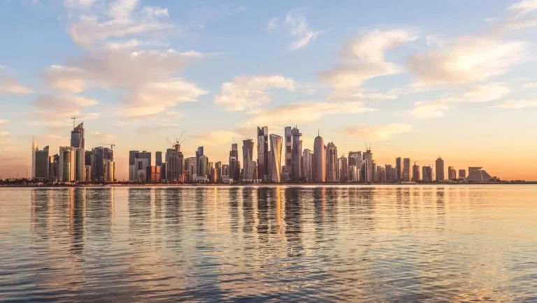 A modern city skyline with tall skyscrapers reflecting in calm water under a partly cloudy sky at sunset, evoking the serenity of a sunrise desert safari.