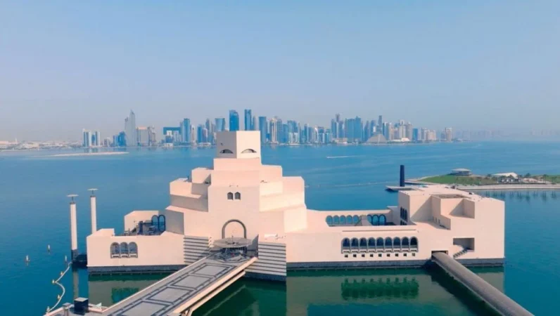 A modern, geometric beige building extends over water with a bridge leading to its entrance; this striking Qatar cultural landmark sits across the bay with the Doha city skyline and tall skyscrapers visible in the distance.