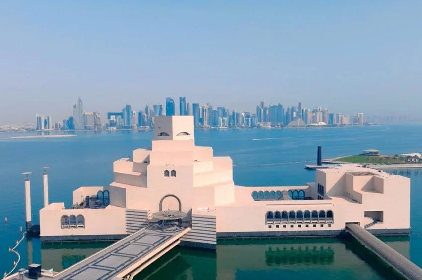 A modern, geometric beige building extends over water with a bridge leading to its entrance; this striking Qatar cultural landmark sits across the bay with the Doha city skyline and tall skyscrapers visible in the distance.