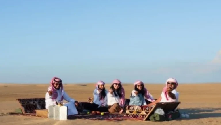 Five people in traditional Middle Eastern attire sit on cushions around a low table, enjoying a Ramadan picnic on a carpet in the Qatari desert, with clear blue skies and endless sand stretching into the distance.