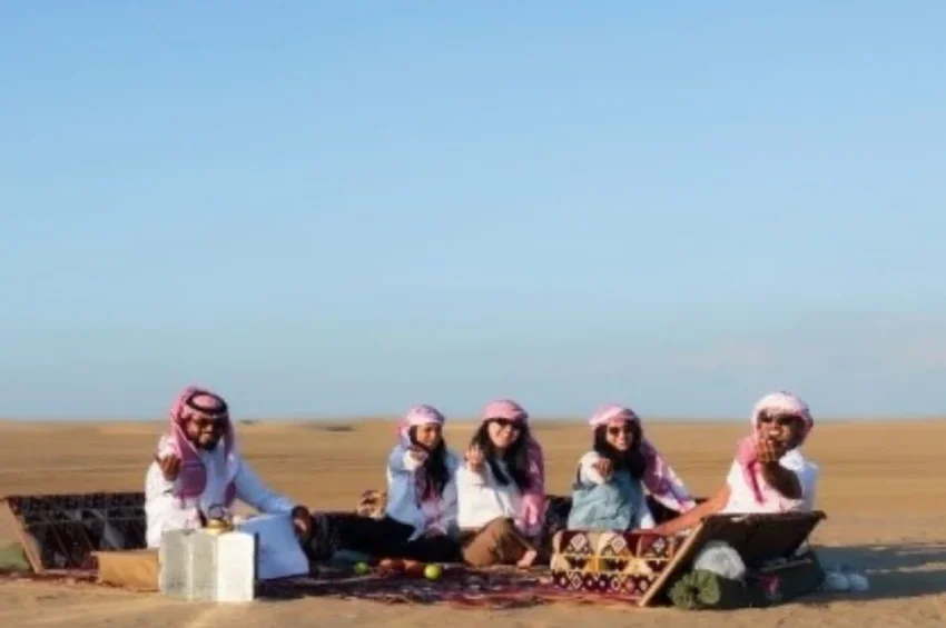 Five people in traditional Middle Eastern attire sit on cushions around a low table, enjoying a Ramadan picnic on a carpet in the Qatari desert, with clear blue skies and endless sand stretching into the distance.