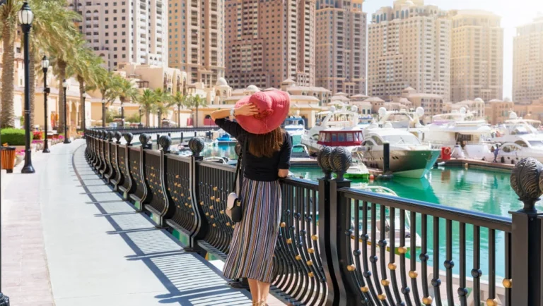 A woman in a striped skirt and pink sunhat walks along a waterfront promenade near the Museum of Islamic Art, with boats, yachts, tall modern buildings, and palm trees in the background on a sunny day.