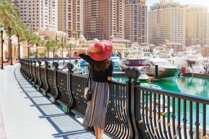 A woman in a striped skirt and pink sunhat walks along a waterfront promenade near the Museum of Islamic Art, with boats, yachts, tall modern buildings, and palm trees in the background on a sunny day.