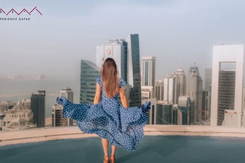 A woman in a blue dress stands on a rooftop, overlooking a city skyline with modern skyscrapers and water below—a perfect scene for female travelers seeking inspiration under the clear sky.
