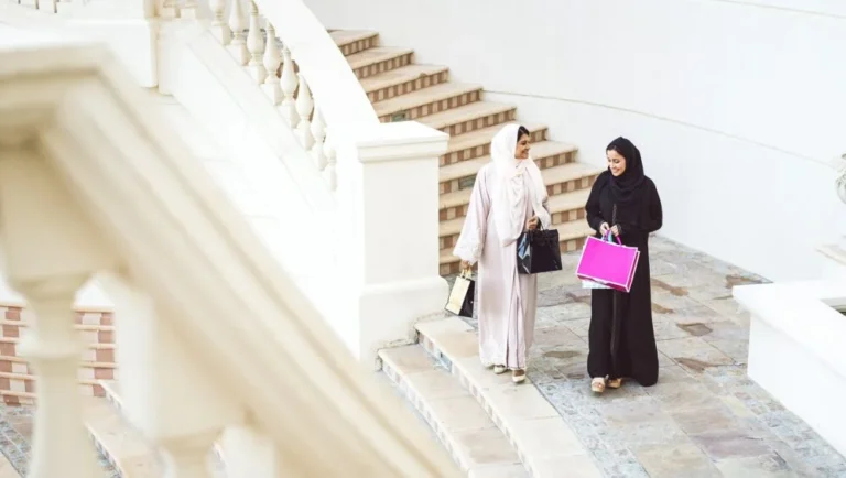 Two female travelers in hijabs, one in light clothing and one in black, walk down outdoor stone steps with shopping bags, chatting as they’re surrounded by white railings and walls.