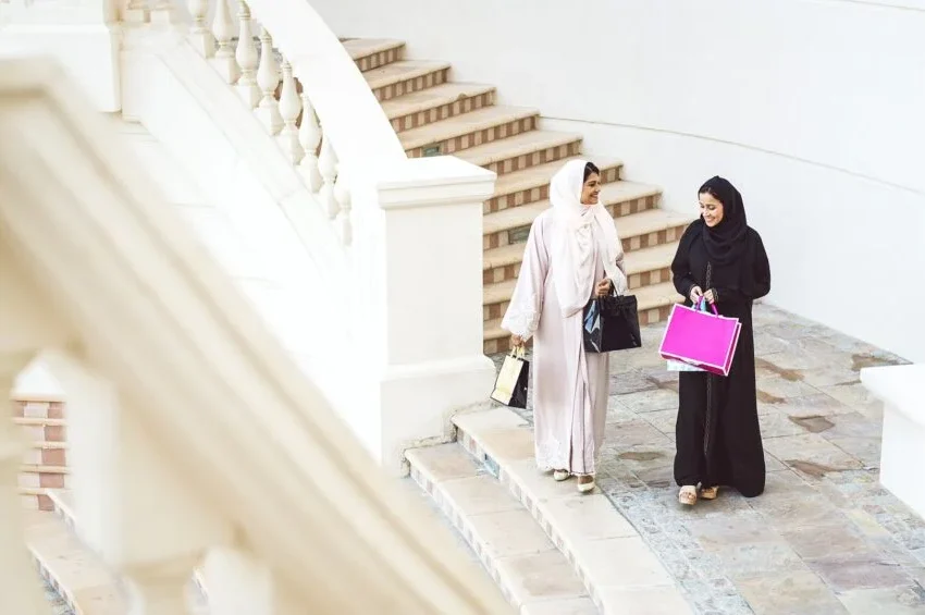 Two female travelers in hijabs, one in light clothing and one in black, walk down outdoor stone steps with shopping bags, chatting as they’re surrounded by white railings and walls.